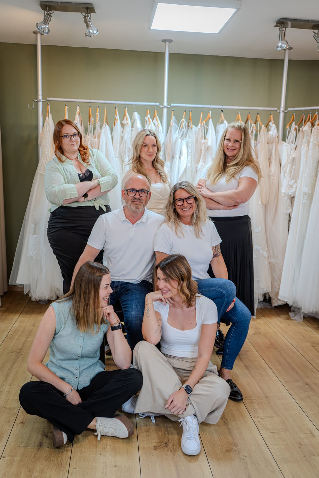 Sieben Mitglieder des La-novia-Teams sitzend und stehend vor Brautkleidern